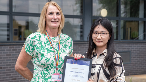 Two people standing in front of a building with large windows; one holding a framed certificate